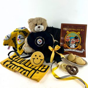 A black-and-gold Steelers-themed baby gift basket featuring a cuddly teddy bear, a taggie blanket, Steelers bib, Terrible Towel, children’s book, italian cookies, and black and gold chocolate covered pretzels, all arranged in a black basket with black and gold ribbon.